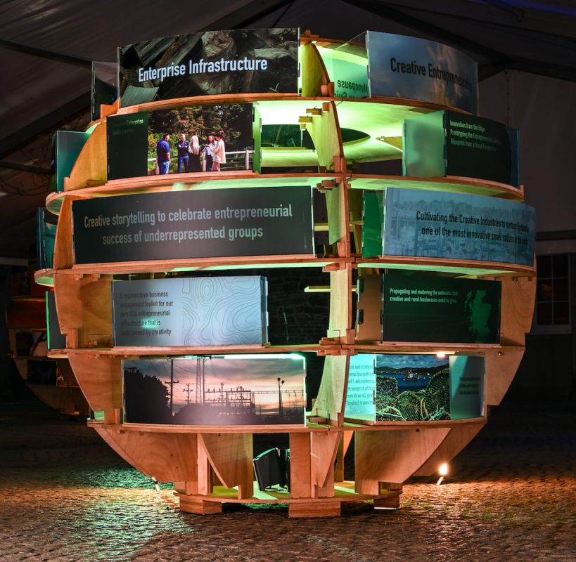 Front view of circular wooden Rural Lab exhibition with green and yellow illuminated content panels.